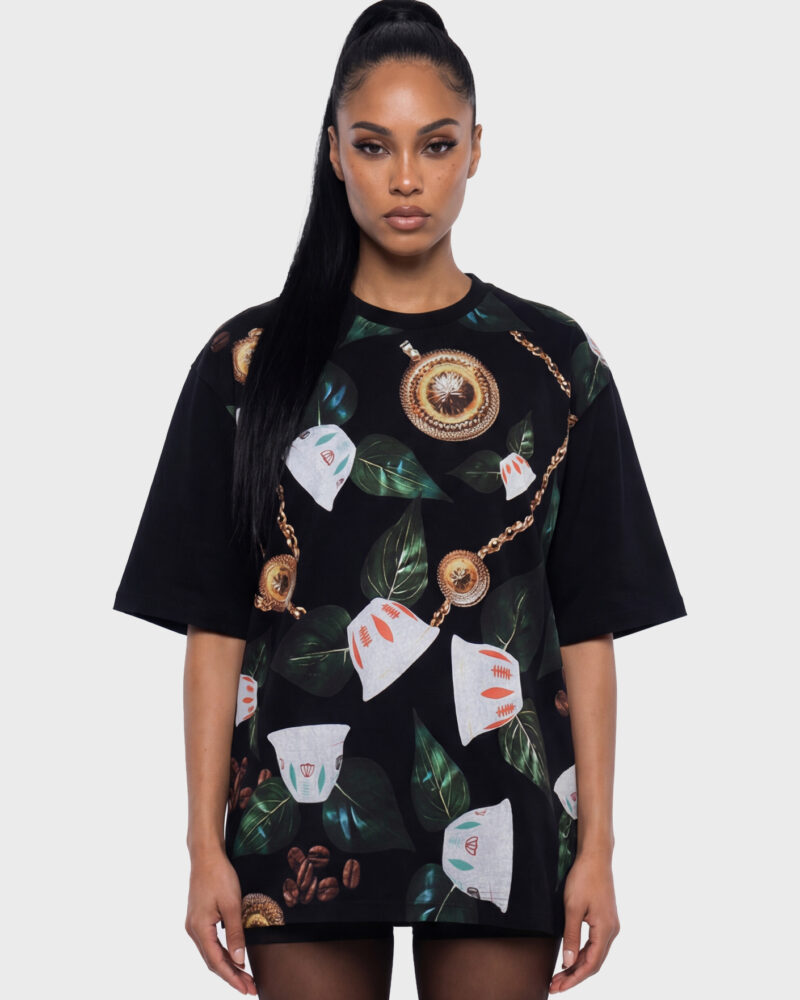 Front view of a woman wearing a black oversized boxy-fit T-shirt with an all-over print inspired by Ethiopia’s coffee ritual, featuring Cini cups, green leaves, roasted coffee beans, and gold medallion accents.