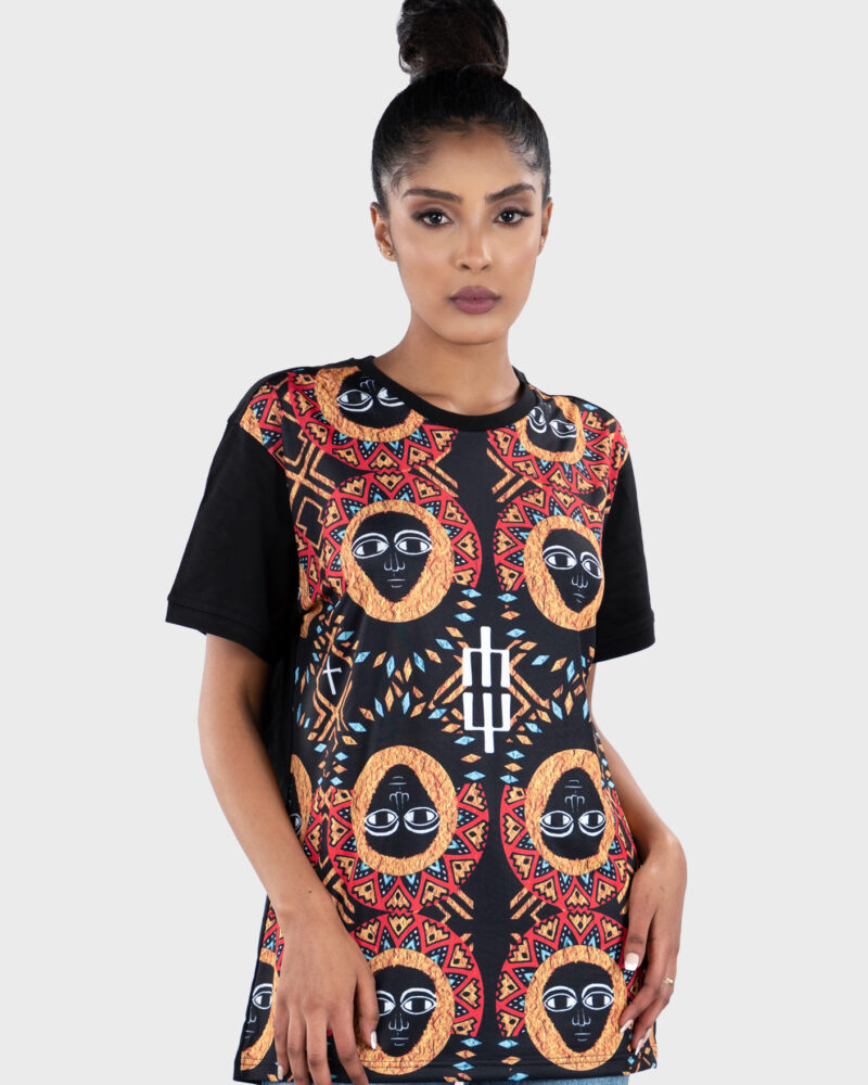 Front view of a woman wearing a black T-shirt with a front-panel Tibeb-inspired print featuring wide-eyed Ethiopian angel faces framed by geometric halos in red, gold, and black tones.