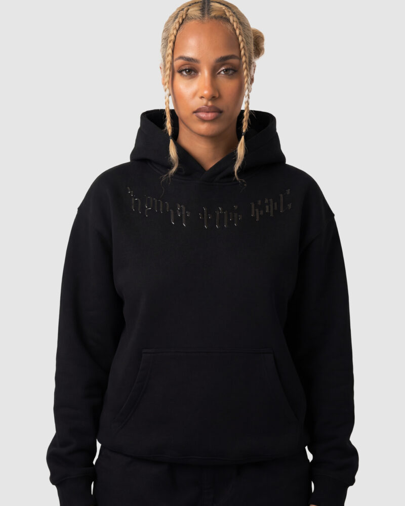 Front view of a woman wearing a heavyweight black hoodie with HF-embossed glossy PU Amharic lettering across the neckline.